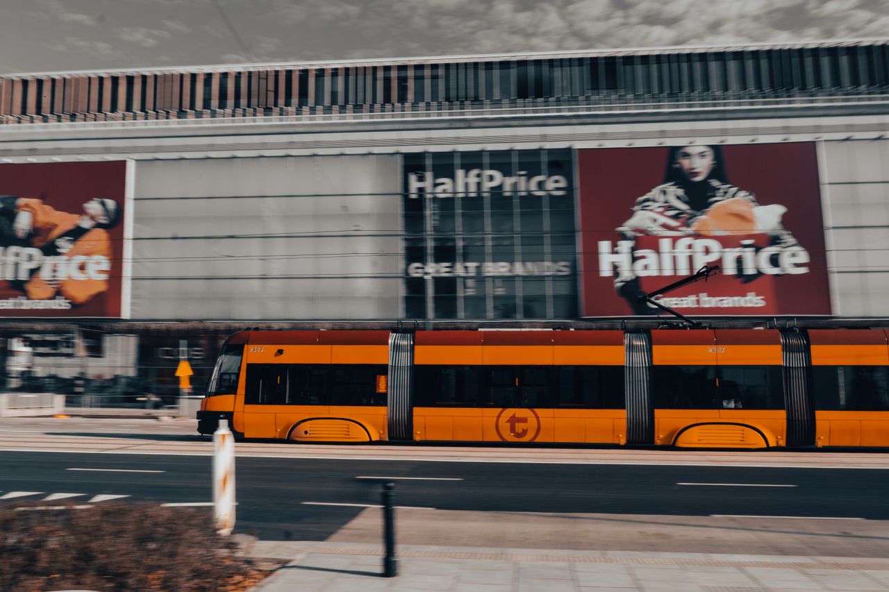 A vibrant yellow tram moving quickly in front of a shopping center in Warsaw, Poland.